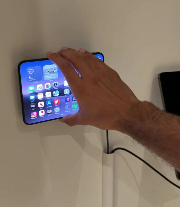 展开的 iPhone Duo 挂在白色直立 MagSafe 充电座上,机身相对支架立柱明显偏转,一只手正按住机身边缘