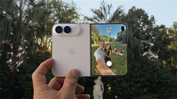 苹果官方 iPhone Duo 宣传图：一只手拿着展开状态的 iPhone Duo，右屏显示取景框内一家人在花园自拍的场景