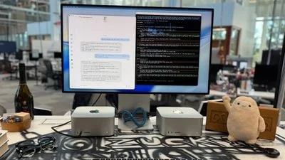 Meta 办公室桌面照片:一台大显示器下方摆着两台银色 Mac Studio,桌面上还有玩偶、墨镜与香槟