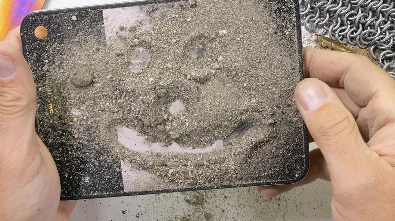 双手托着折叠状态的 iPhone Duo,机背整面铺满沙粒并被画成一个笑脸,右上角露出一块金属环链清洁块,背景是浅色桌面