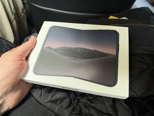 手持白色 iPhone Duo 包装盒，盒面印着展开状态的机身与山景壁纸，背景是黑色行李箱和外套