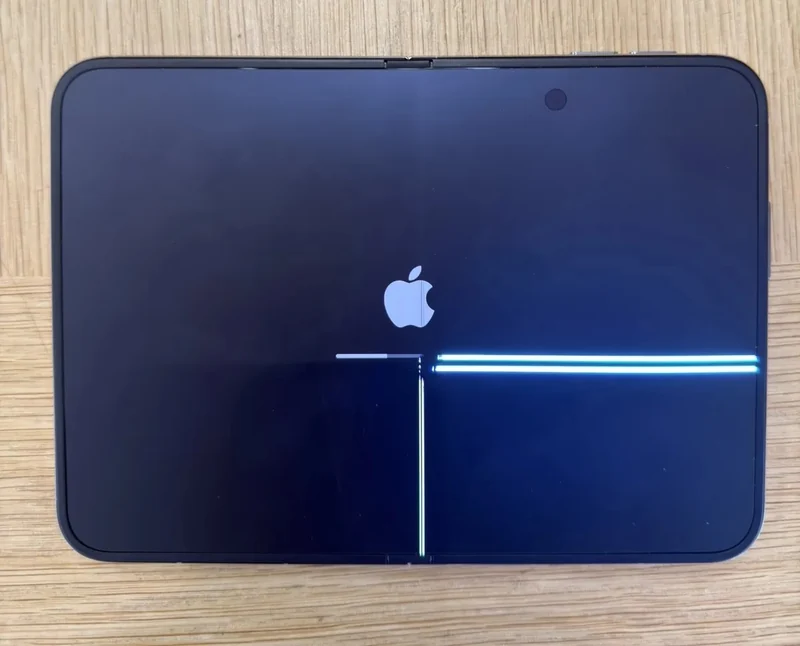 一台展开平放的 iPhone Duo,内屏中央沿折痕出现竖直亮线,右侧另有一条横贯亮带,屏幕显示 Apple 标志