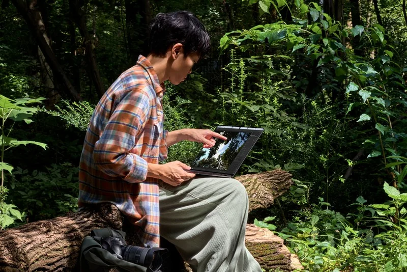 一个人坐在森林里的枯木上,手指点在 MacBook Pro 屏幕上,四周是茂密的绿色植物与透过树叶的阳光,机身旁边放着相机