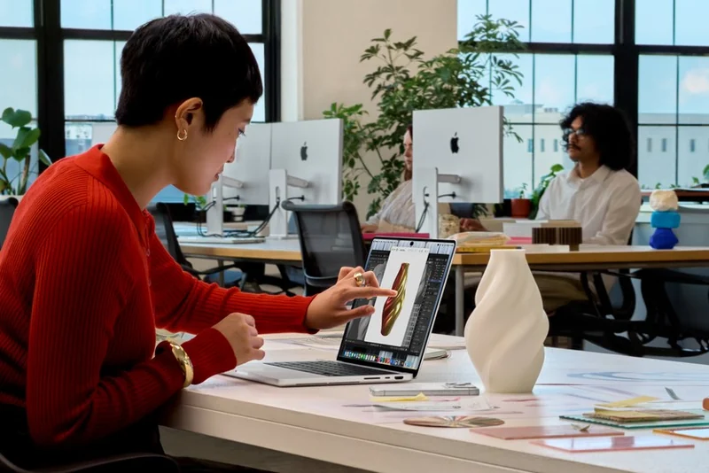 一位穿红色毛衣的短发女性坐在明亮的办公室里,右手食指正点在 MacBook Pro 屏幕上,屏幕显示一张金色螺旋状的立体渲染图,背景有其他 iMac 与同事