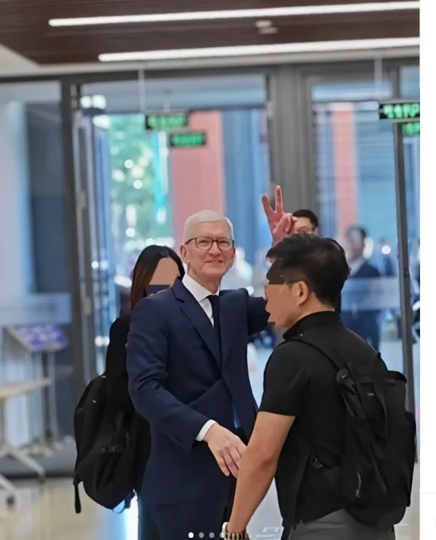 库克穿深色西装在清华大学经济管理学院楼内与学生打招呼,右手举起比出胜利手势