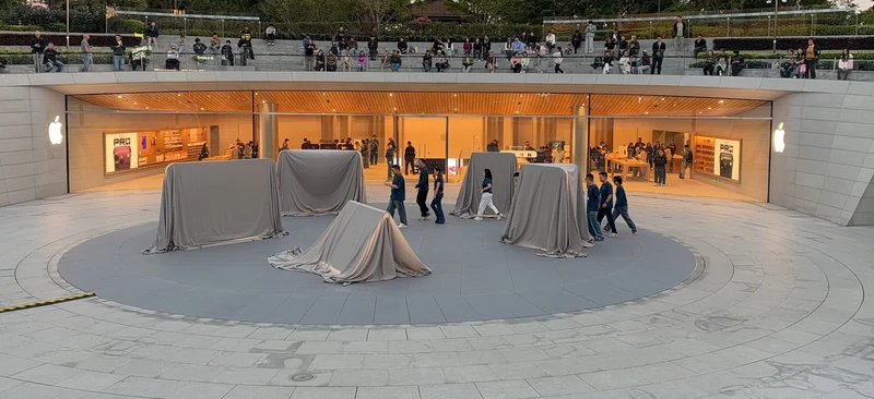 上海静安 Apple Store 广场，几座雕塑仍被银灰色布罩覆盖，旁边有身穿深色服装的工作人员在布置，台阶与围栏边站着围观人群