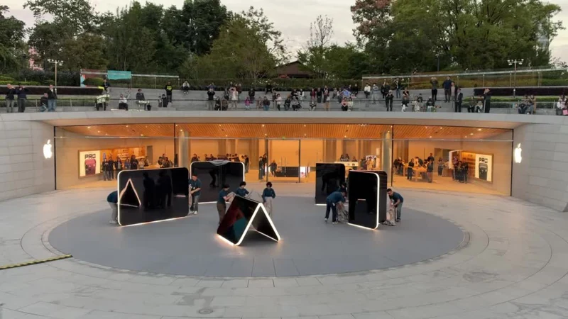 上海静安 Apple Store 下沉广场，多座镜面 iPhone Duo 雕塑立在圆形地面上，其中一座折成三角形态并亮起彩色画面，围栏外与台阶上站满围观人群